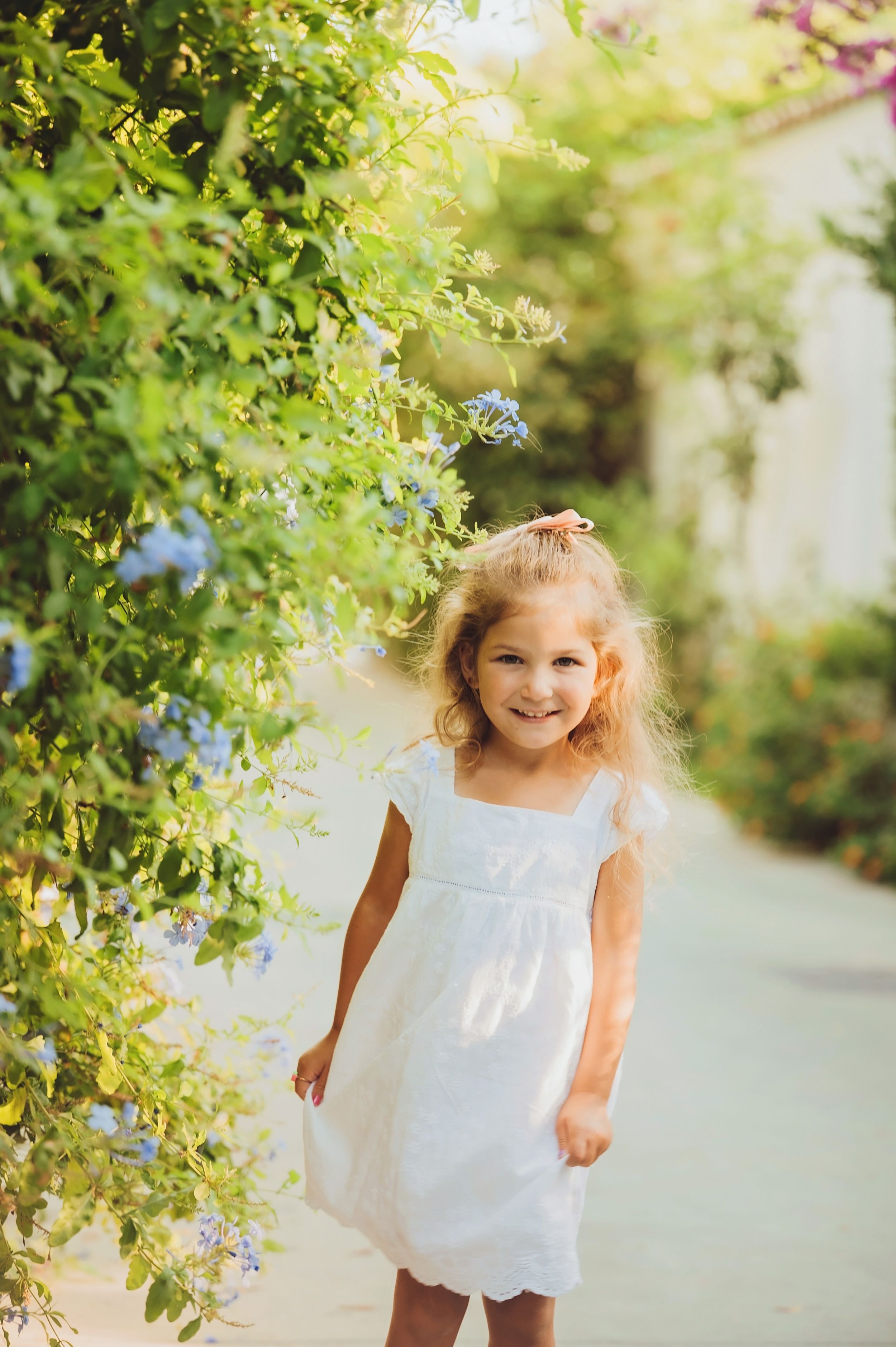 A child in a white summer dress stands near flowering bushes on a garden path.