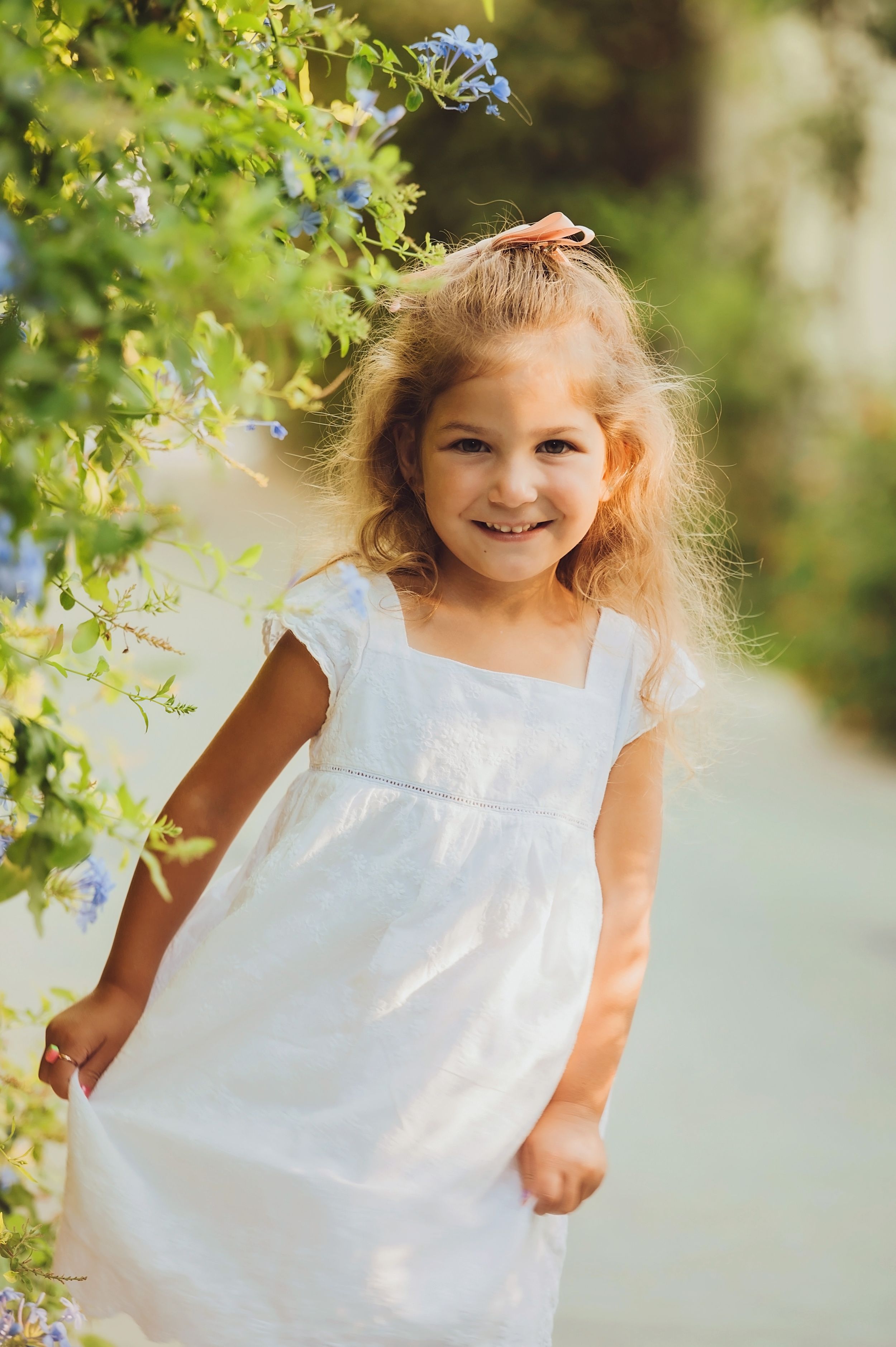 A young child in a white dress stands by blooming blue flowers in a garden setting.