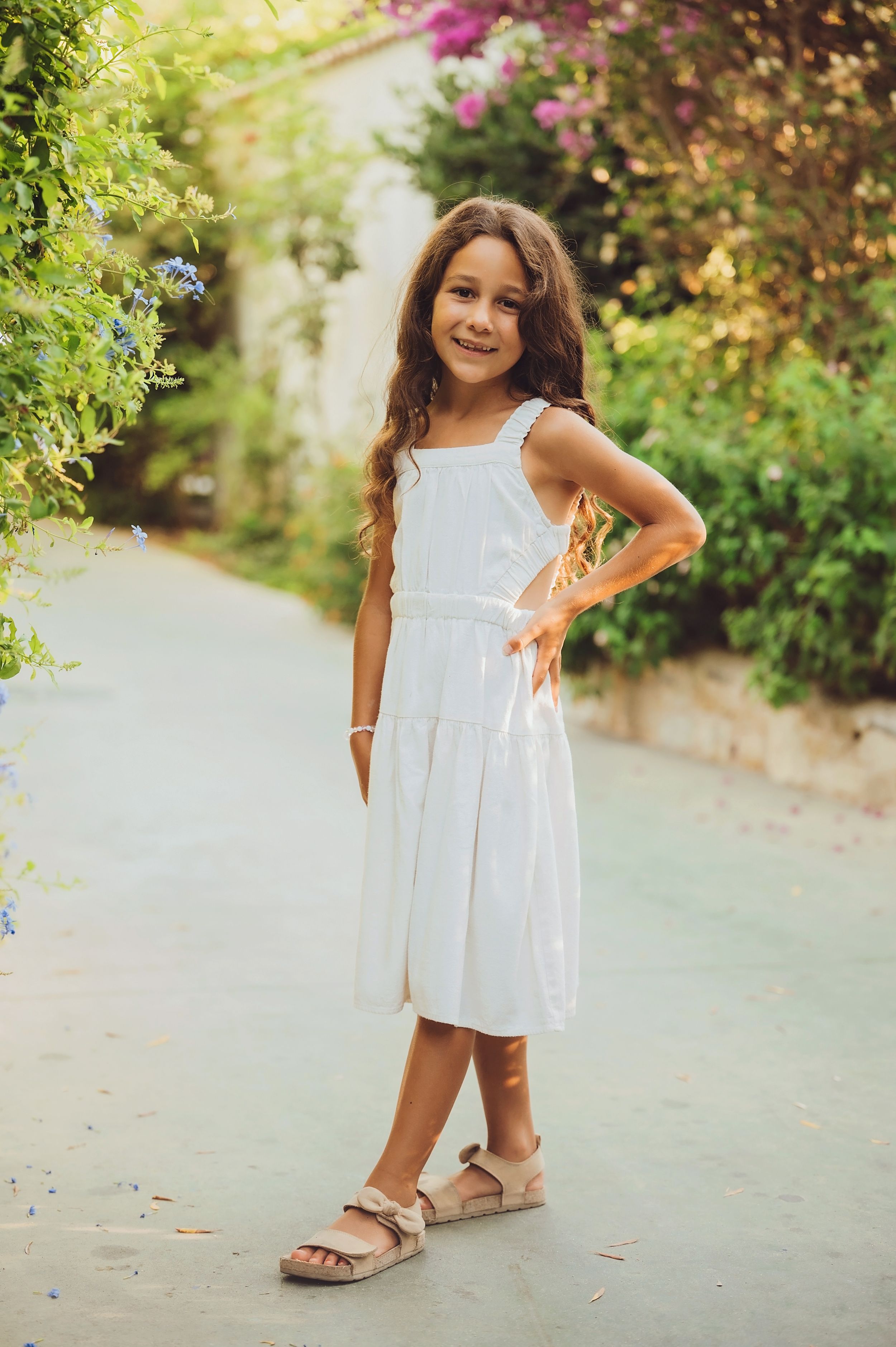 Young child in a white summer dress poses playfully on a garden path during golden hour.