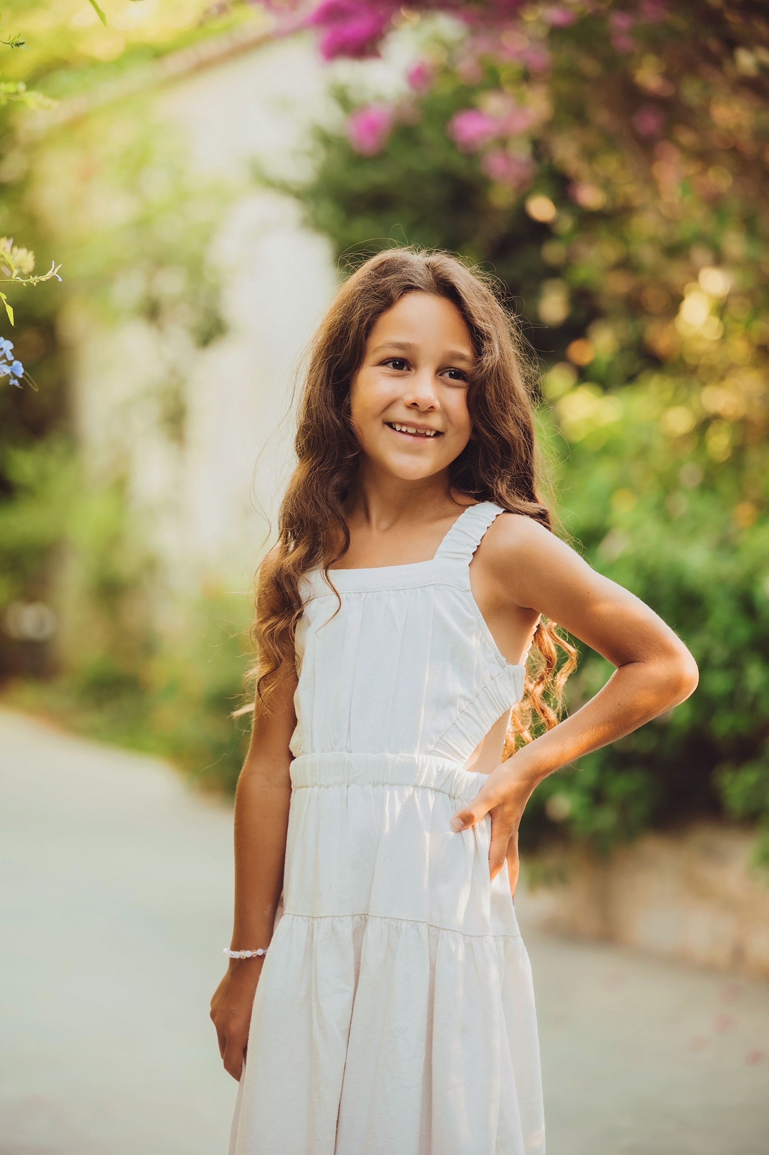 Young girl in a white summer dress poses in a garden setting with blooming pink flowers and greenery.