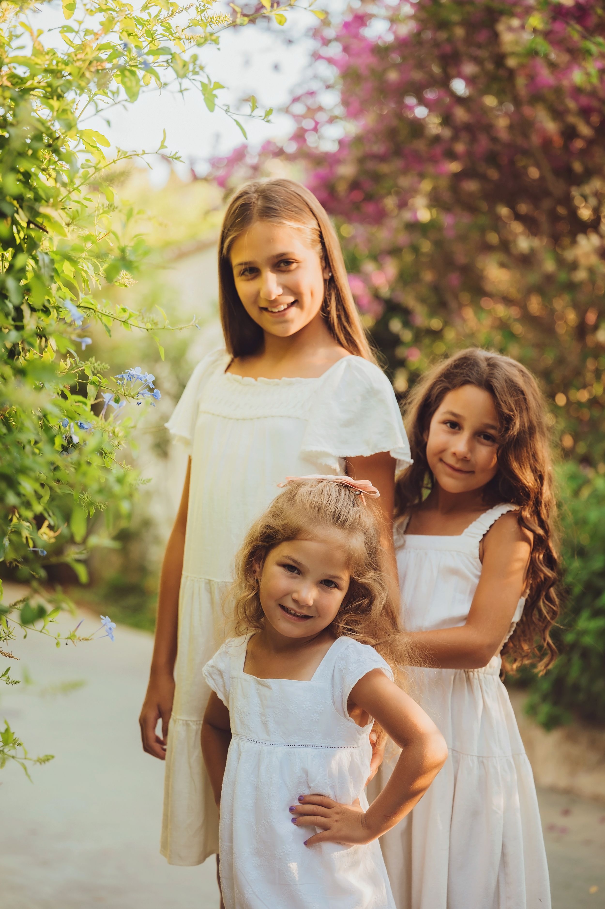 Two children in white dresses pose together in a garden with blooming pink flowers and greenery.