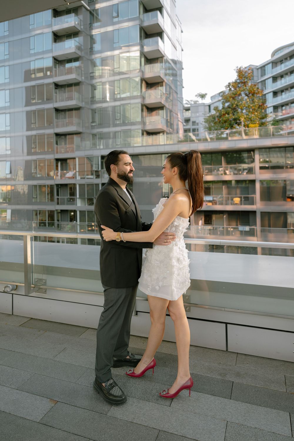 Couple in formal attire embracing on urban balcony, man in black suit, woman in white dress with red heels.