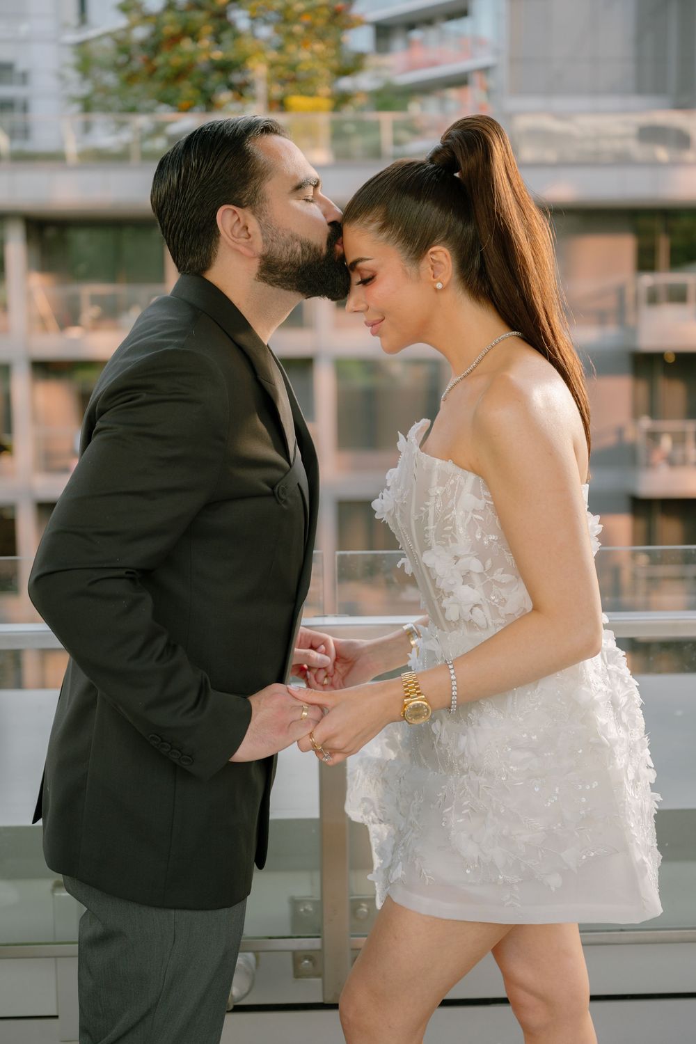 A couple shares a romantic kiss on a rooftop, man in dark suit, woman in white mini dress holding a champagne glass.