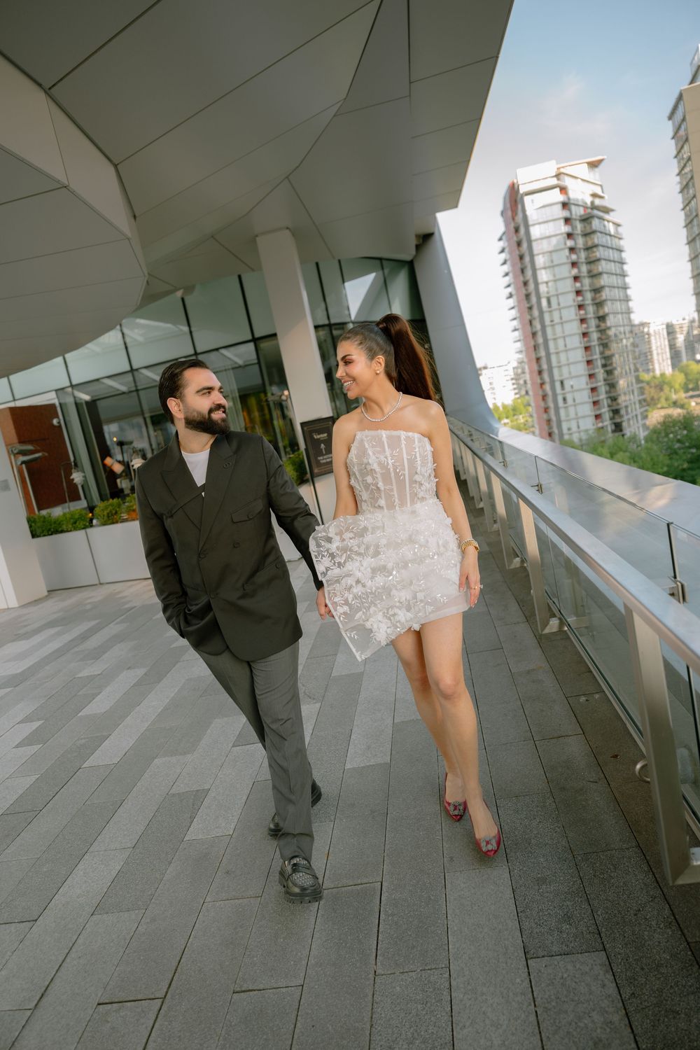 Couple walking hand in hand on a modern urban terrace, woman in white mini dress, man in olive suit, city skyline background.
