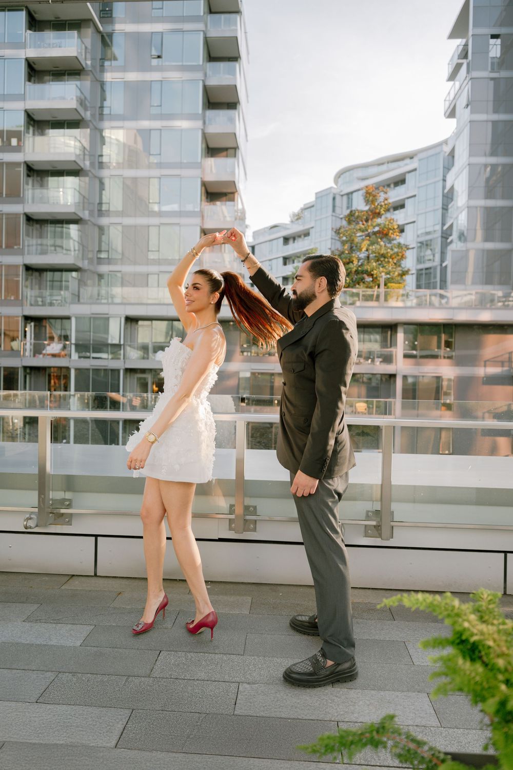 Couple dancing on urban terrace, woman in white dress and red heels, man in dark suit, modern glass buildings backdrop.