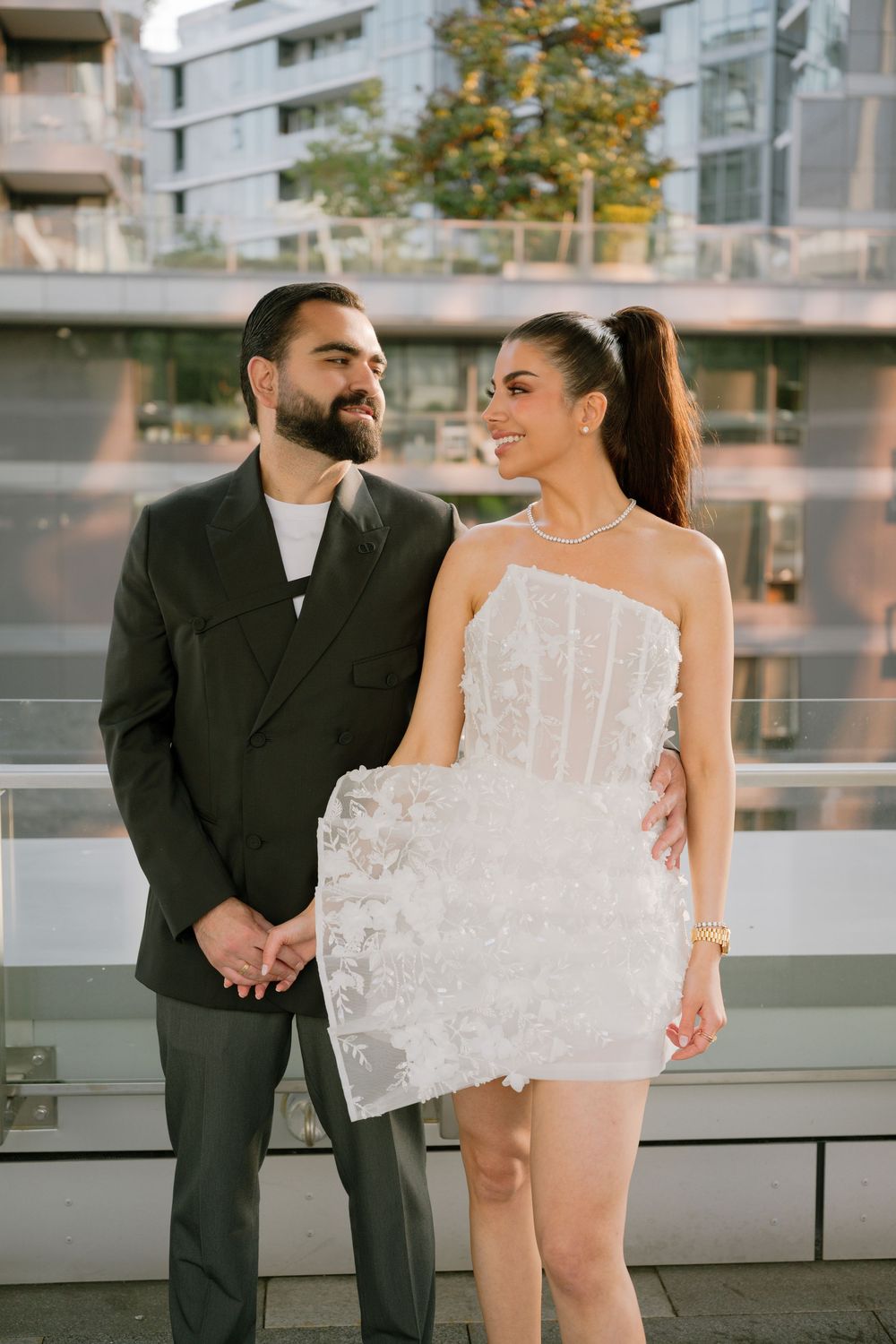 Elegant couple posing outdoors, man in dark suit and woman in white lace mini dress with ponytail, urban backdrop.