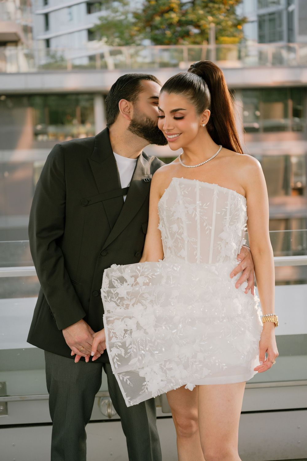A man in a black suit kisses a woman in a white lace mini dress on the cheek as they hold hands outdoors.