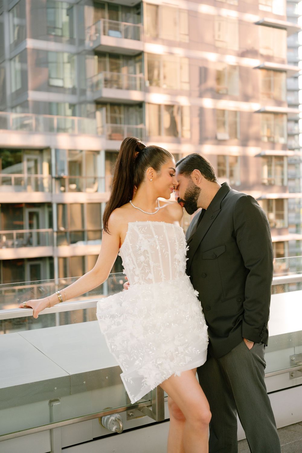 Couple in elegant attire almost kissing on urban rooftop, woman in white mini dress, man in black suit.