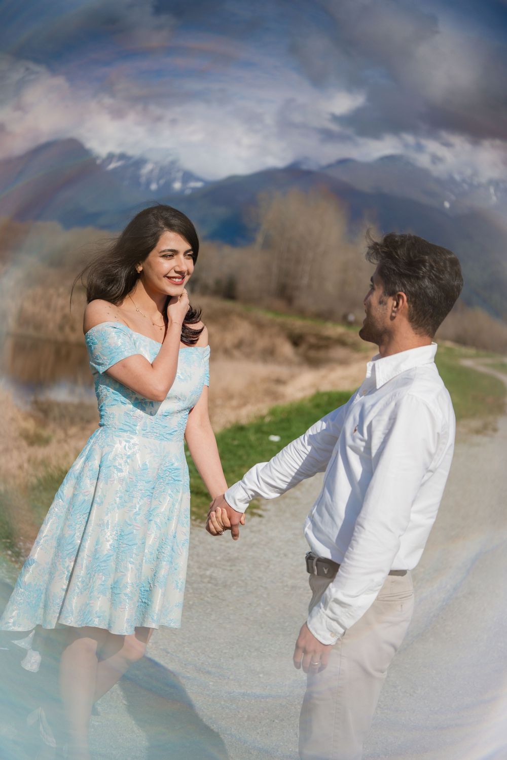 A couple holds hands on a scenic mountain path, the woman in a light blue dress smiling joyfully at her partner.