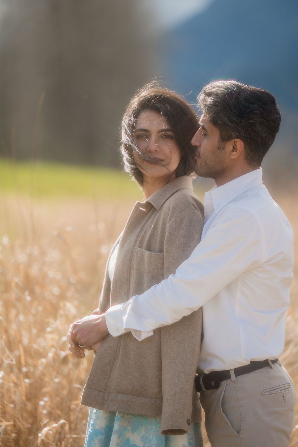 Couple embracing in golden field, man in white shirt holds woman in tan jacket, soft bokeh background.