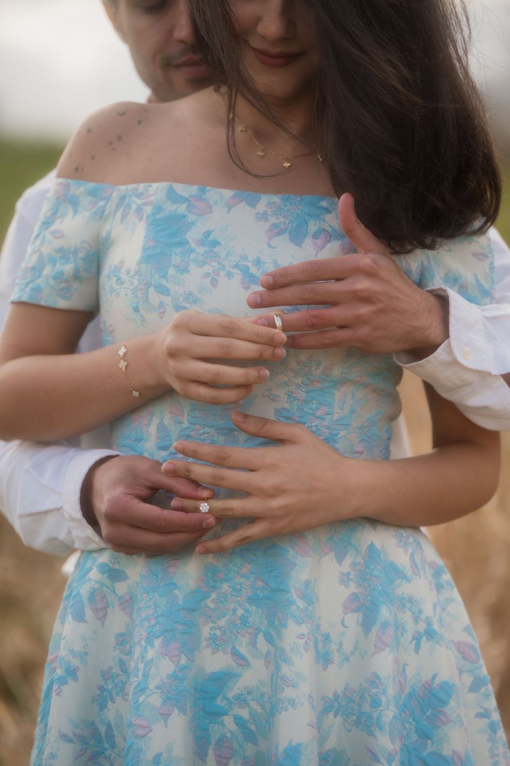 Couple embracing in a field, woman in blue floral off-shoulder dress with engagement ring, man in white shirt holding her.