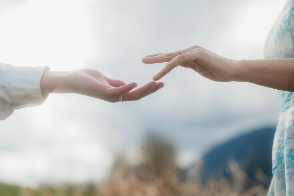 Two people reaching toward each other with outstretched hands in a serene outdoor field with mountains in the background.