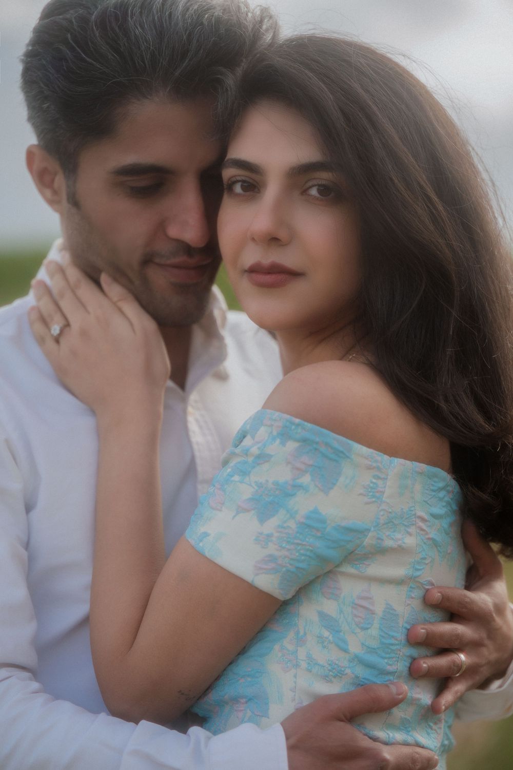 A couple in a romantic embrace, woman in blue floral off-shoulder dress, man in white shirt, outdoors.