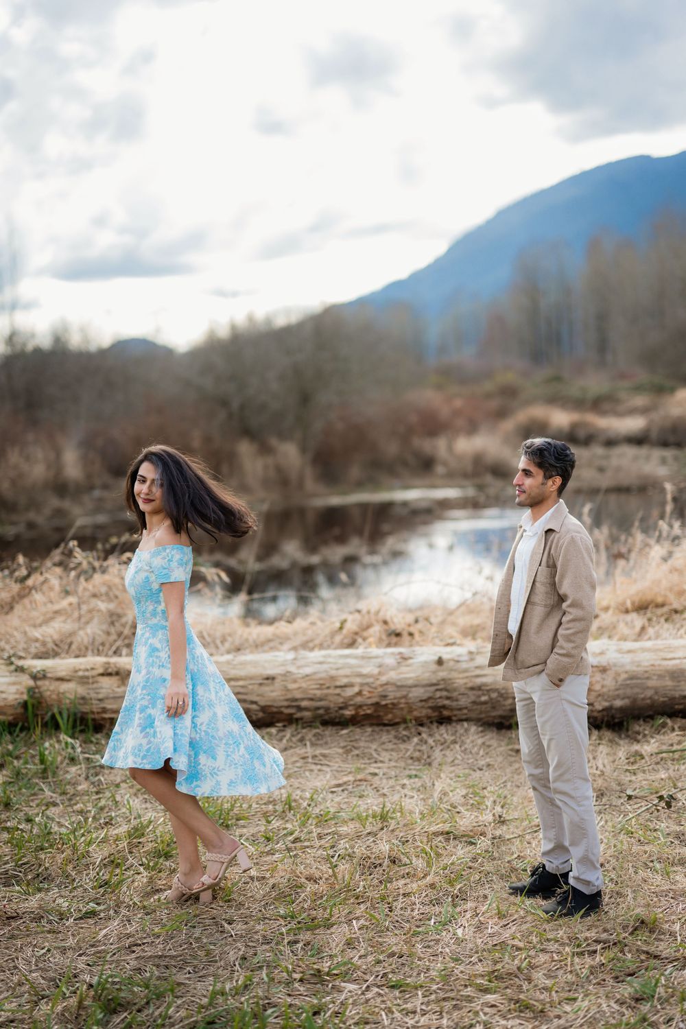Couple posing outdoors near a mountain lake, woman in blue floral dress and man in beige jacket on a cloudy day.