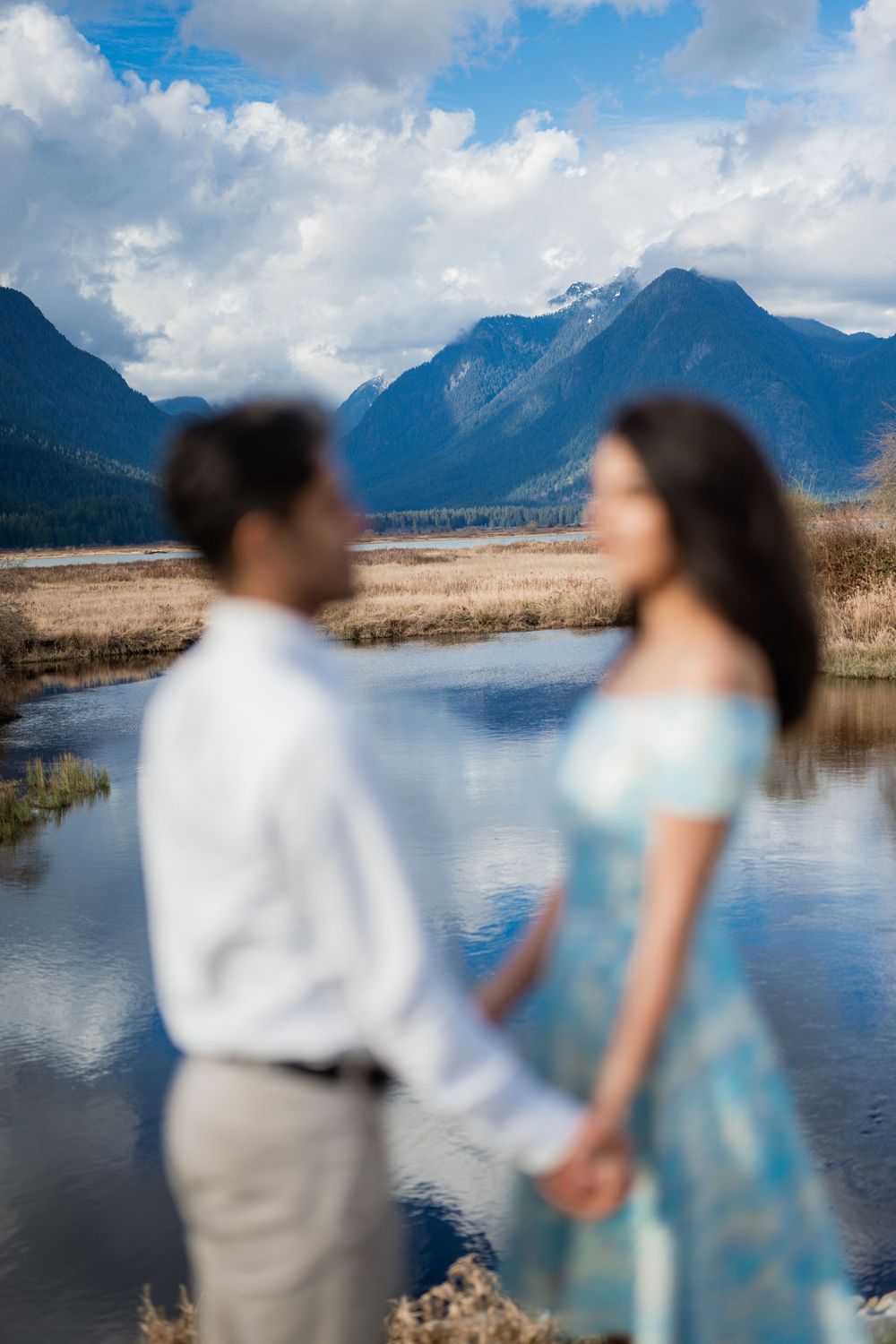 Couple holding hands with blurred foreground, mountain lake and cloudy sky backdrop in a romantic outdoor engagement photo.