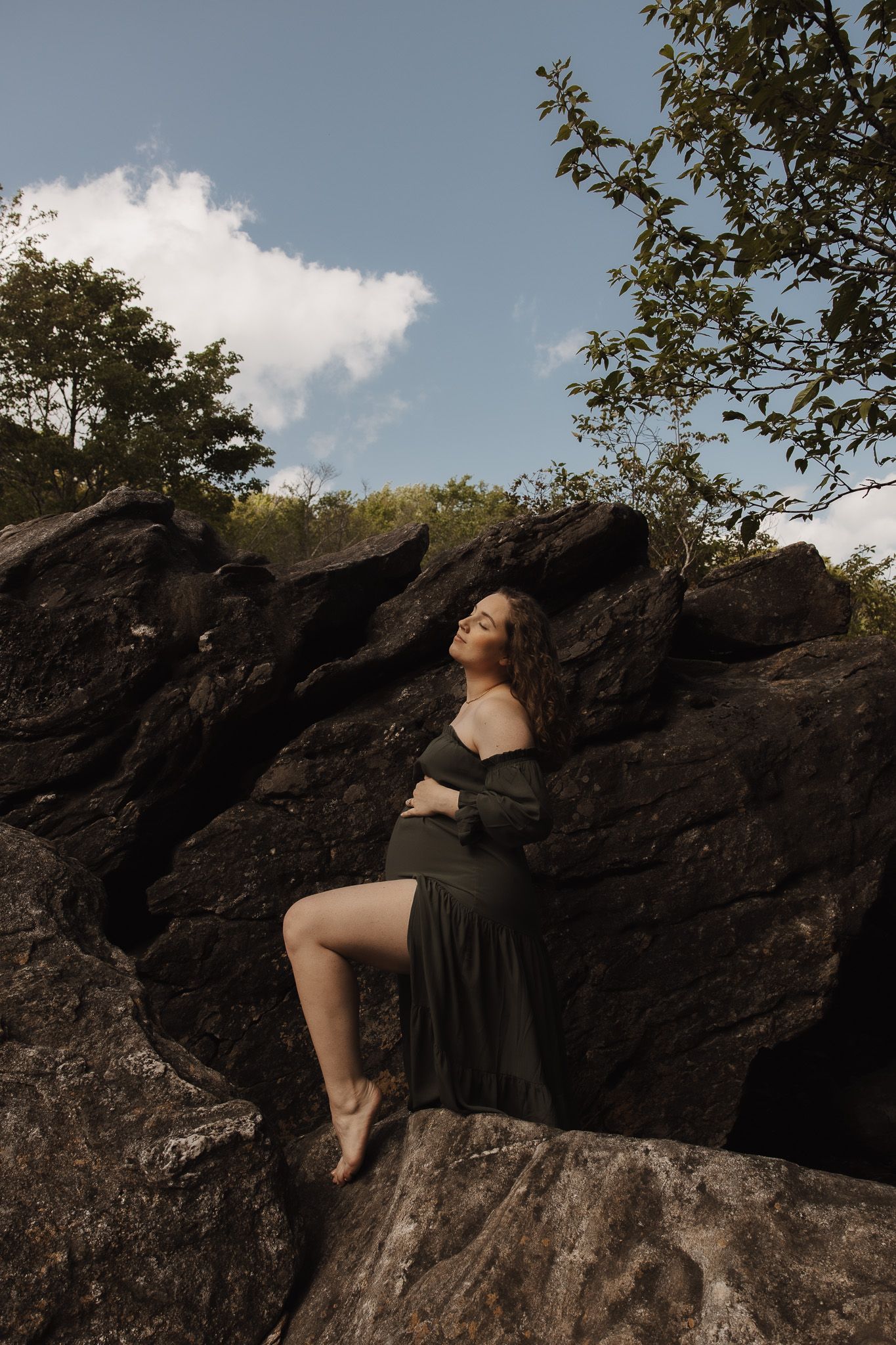 A person in dark clothing reclines on a dramatic rocky outcrop against a blue sky with scattered clouds.