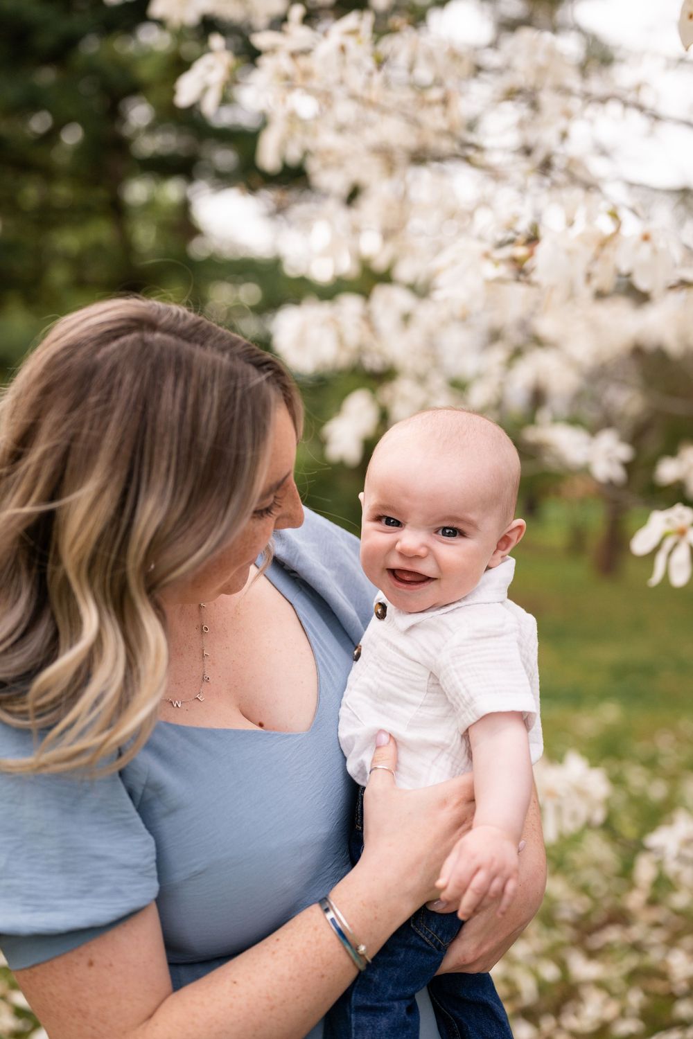 A mother holds her smiling baby outdoors among blooming cherry blossom trees in springtime.