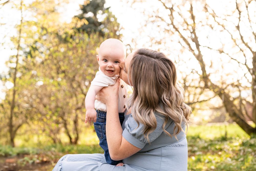 A parent and baby share a tender outdoor moment in a sunlit garden with autumn trees in the background.