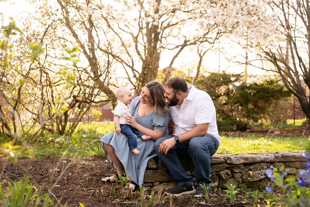 A family sits together on a rustic wooden bench surrounded by blooming spring trees in a natural outdoor setting.