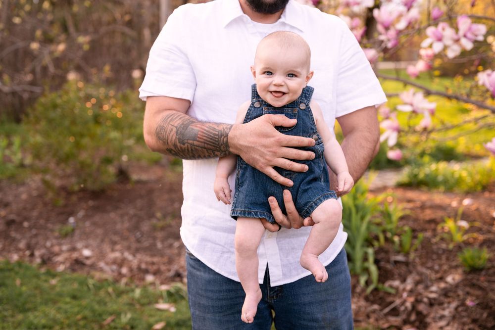 A baby wearing denim overalls is held by someone in a white shirt outdoors with pink blossoms in the background.
