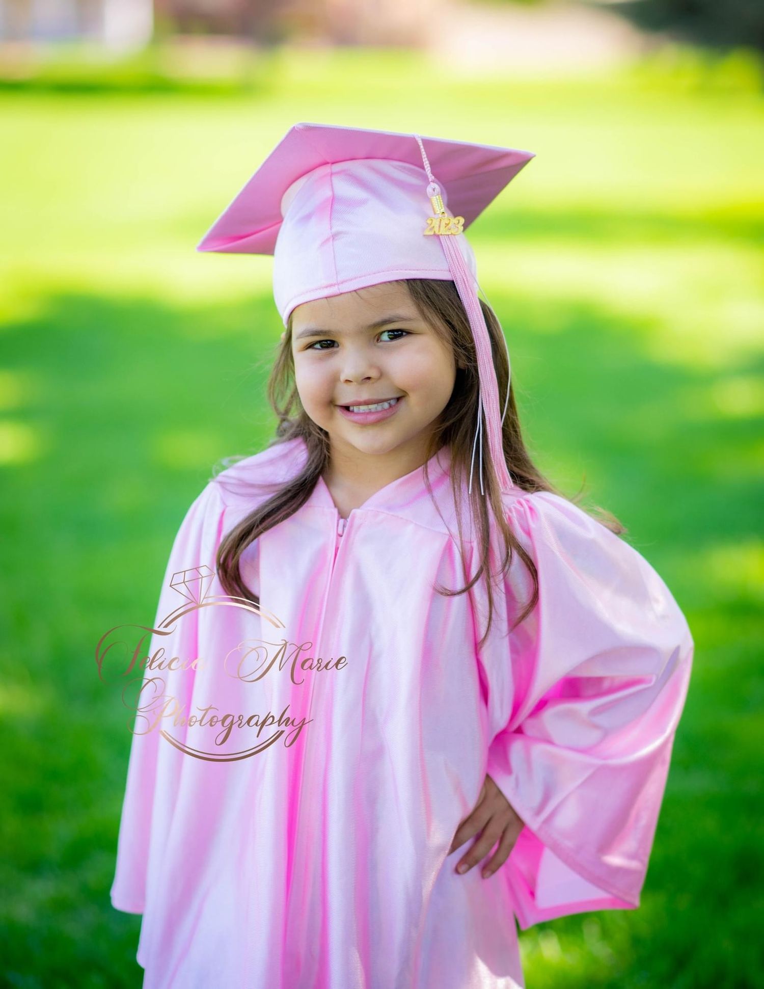 A child wearing a pink graduation cap and gown smiles while posing outdoors.