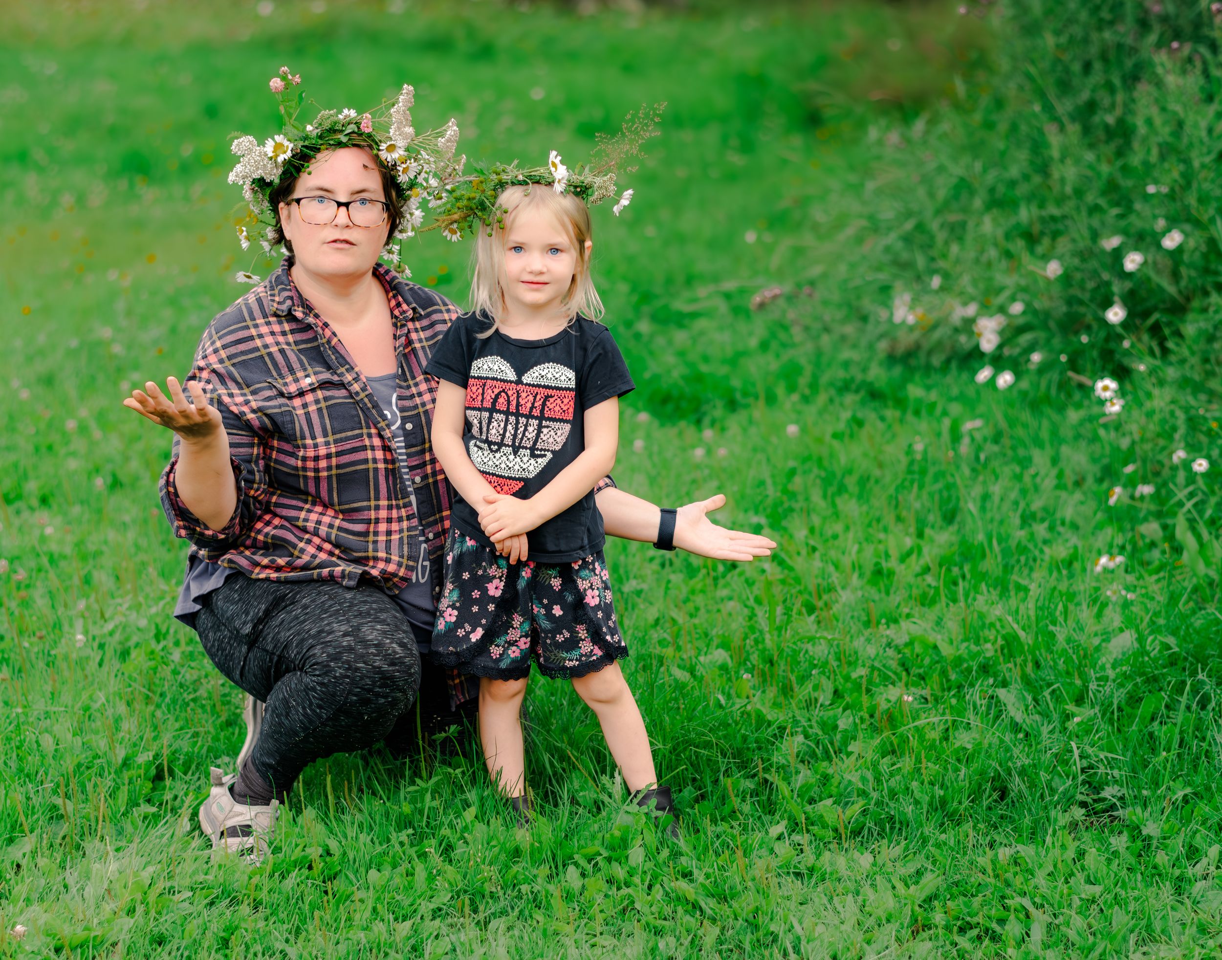 A person and child sit together on a lush green hillside scattered with small white wildflowers on a sunny day.
