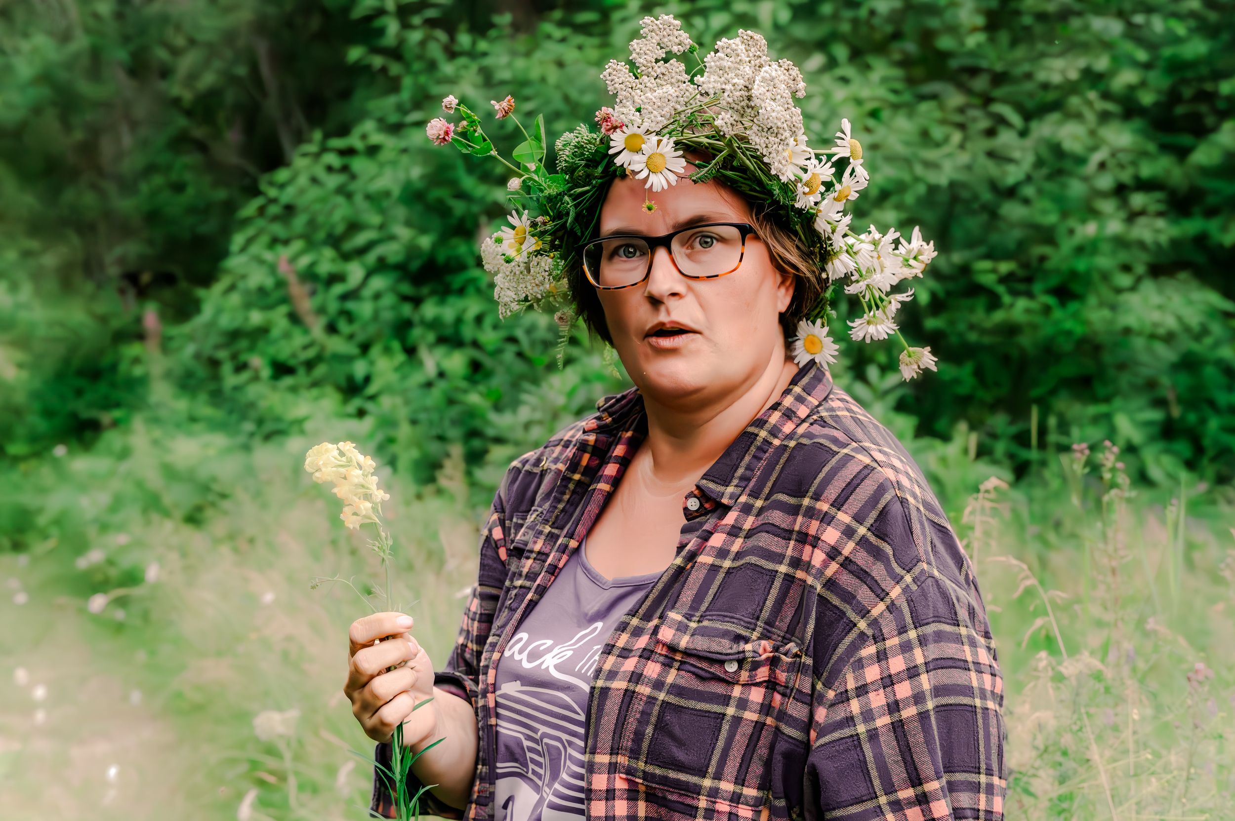 A person wearing glasses and a plaid shirt poses outdoors with white flowers in their hair in a natural setting.