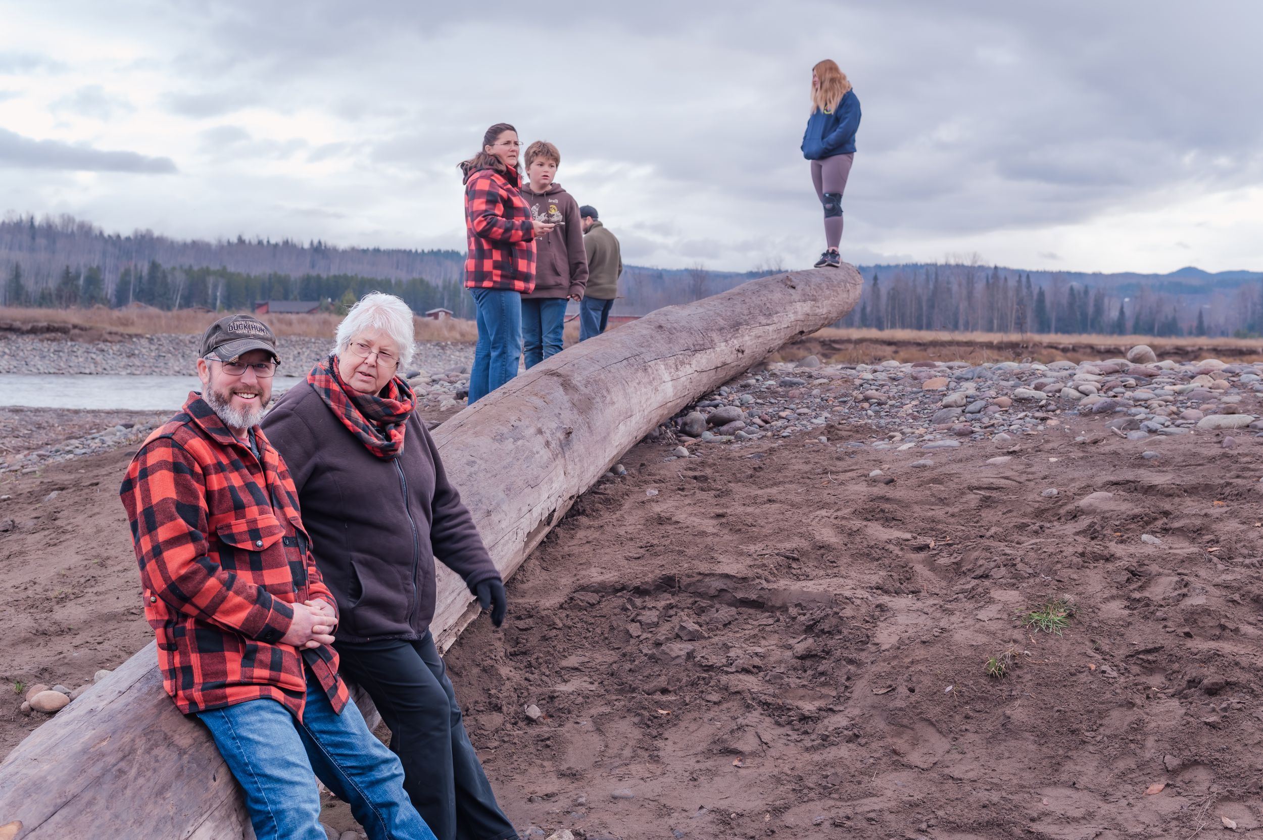 Several people explore a massive fallen log at Mount St. Helens National Volcanic Monument on a cloudy day.
