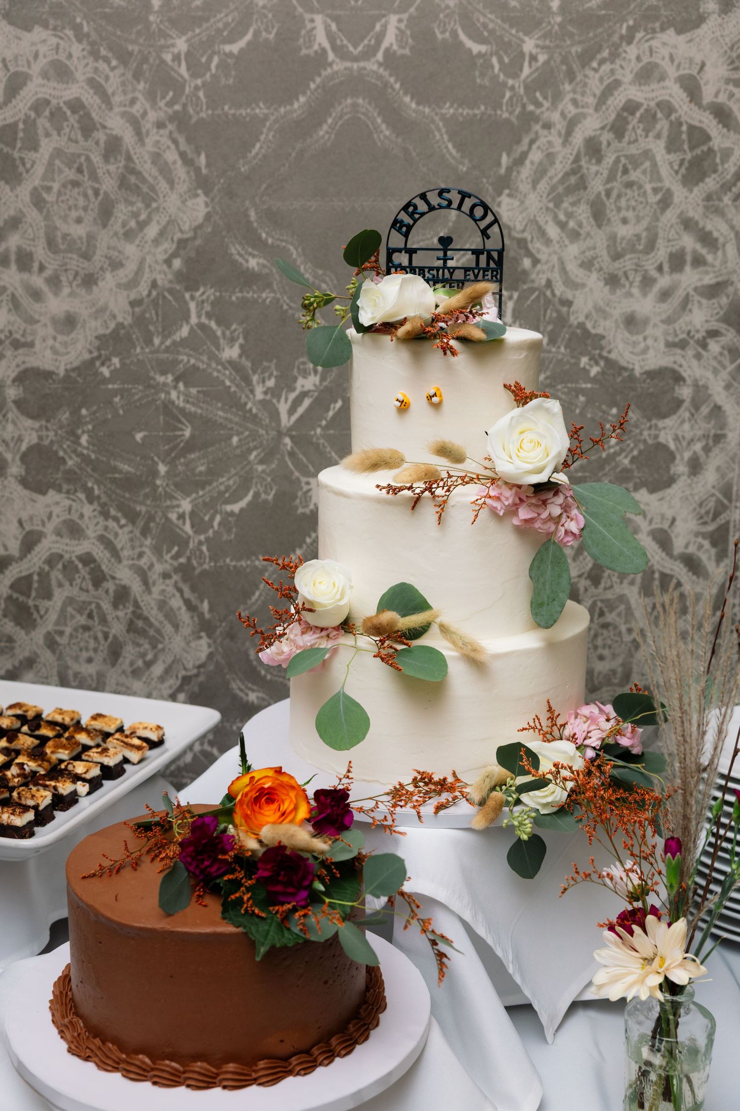 Elegant three-tier white wedding cake with floral decorations and eucalyptus leaves displayed next to chocolate cake with fresh flowers.