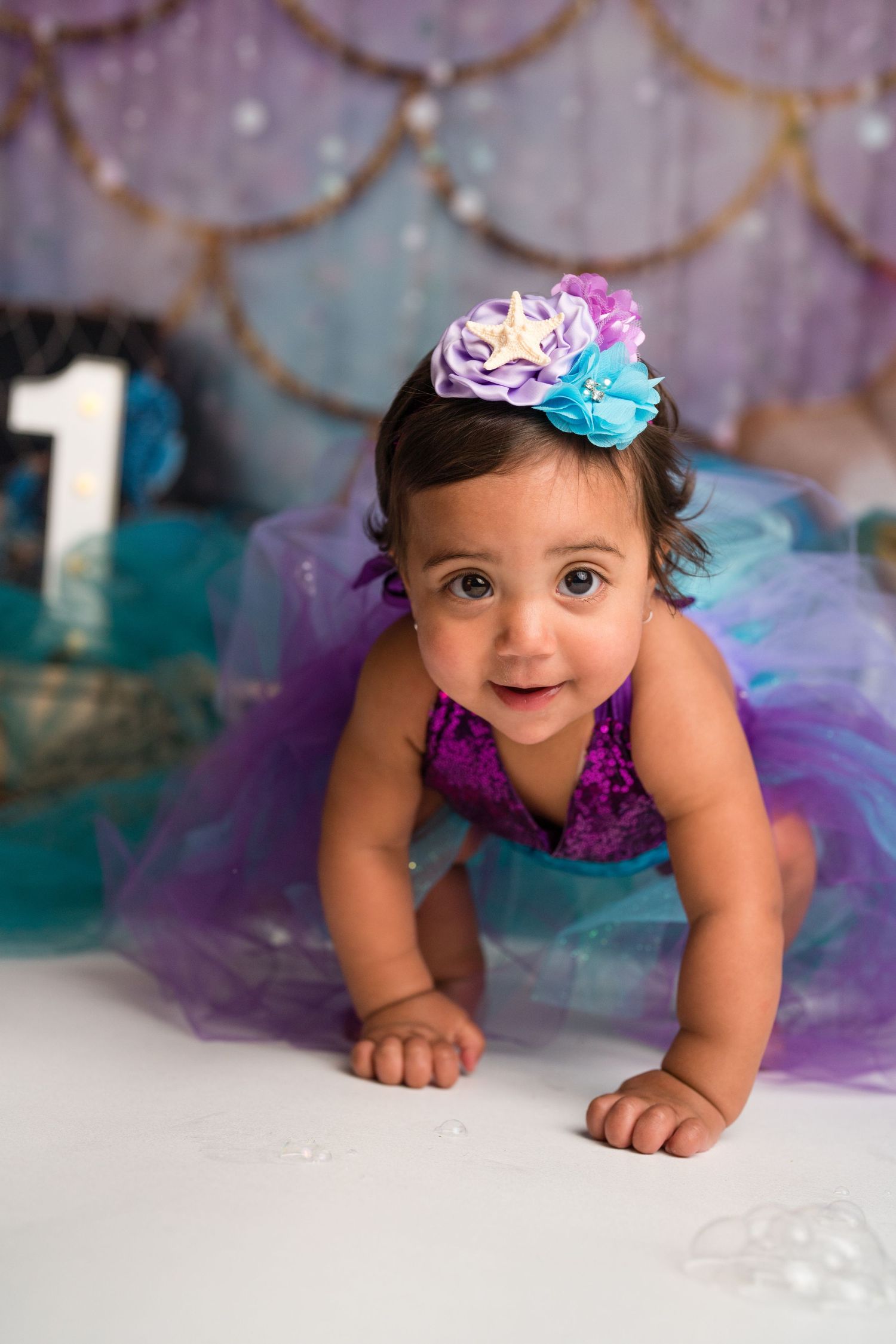 Baby wearing purple tutu and floral headband crawls on white backdrop with sparkly decorations in background.