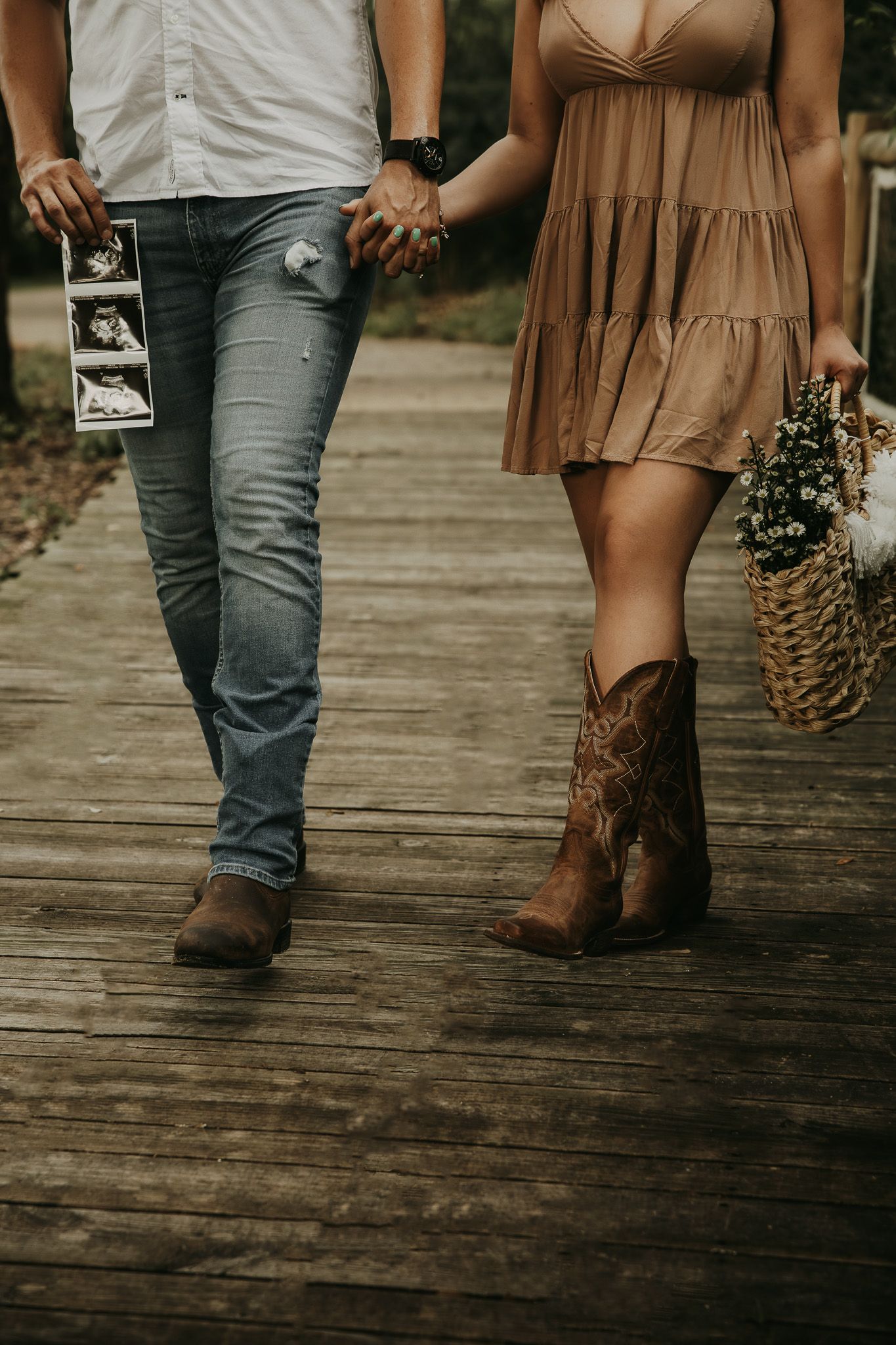 Couple holding hands and ultrasound photo walking on wooden boardwalk in casual western attire.