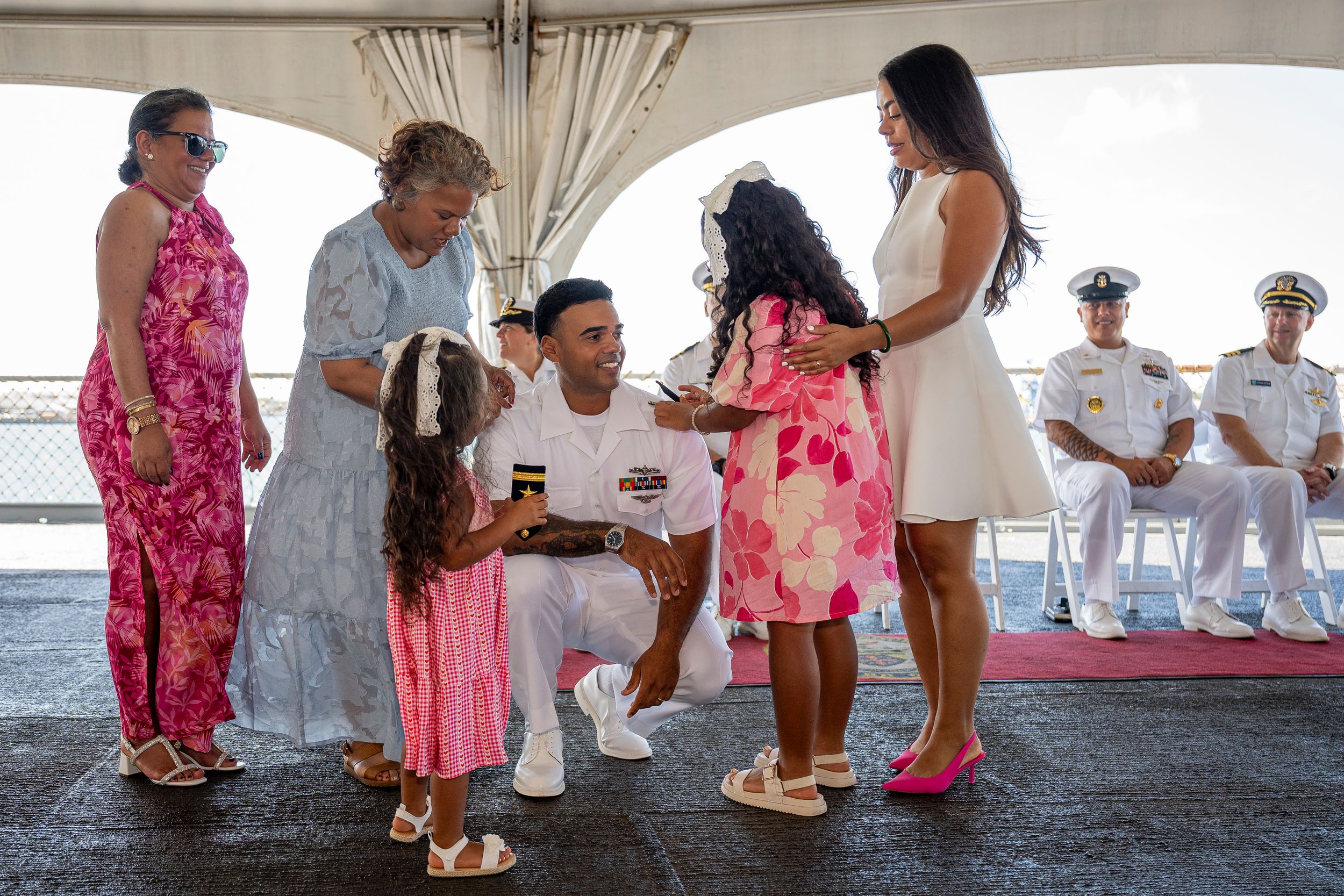 Navy sailor reunites with family on deck, kneeling to greet children in a heartfelt homecoming ceremony.