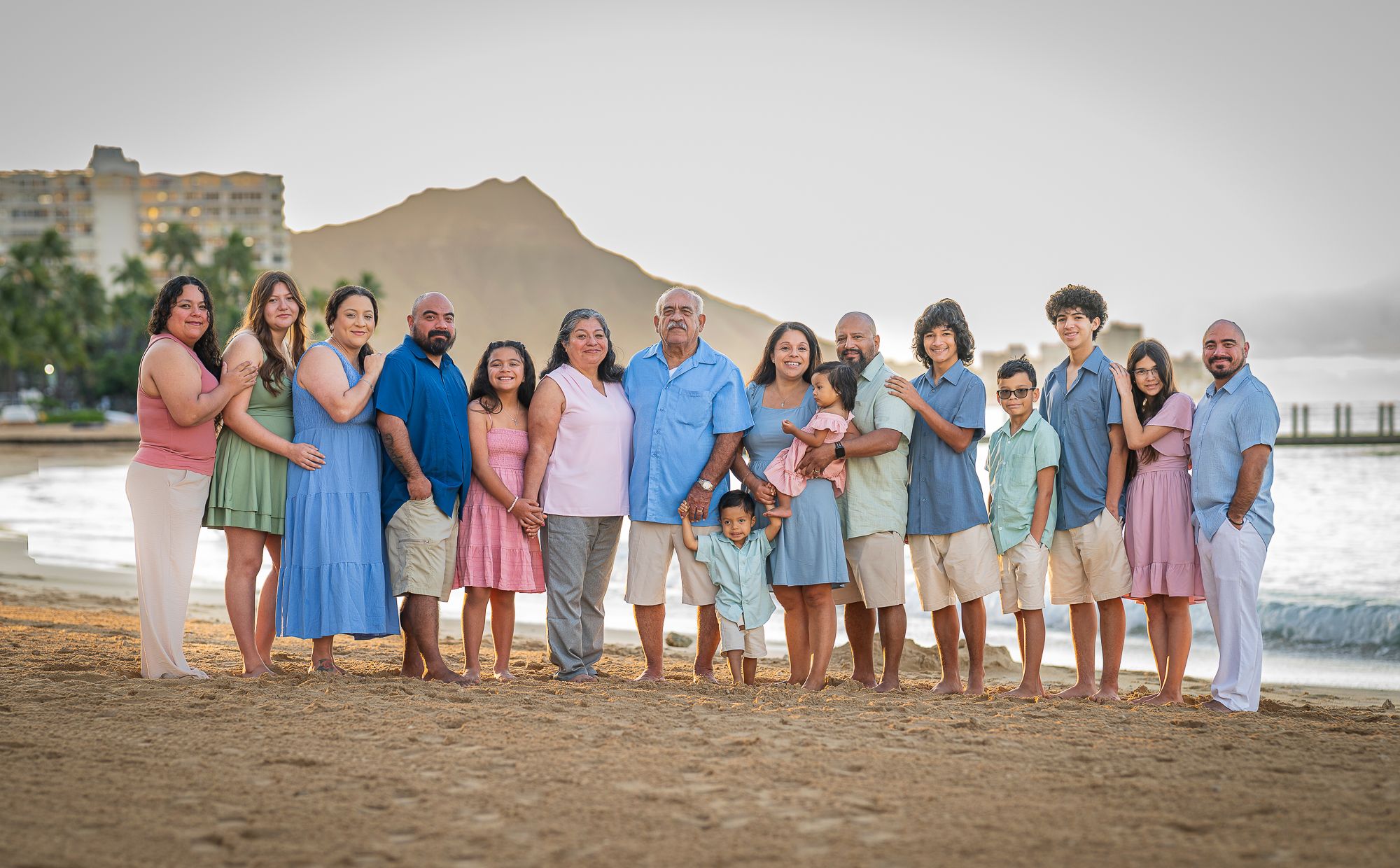 Large family group wearing coordinated pastel outfits posing on Waikiki beach at sunrise.