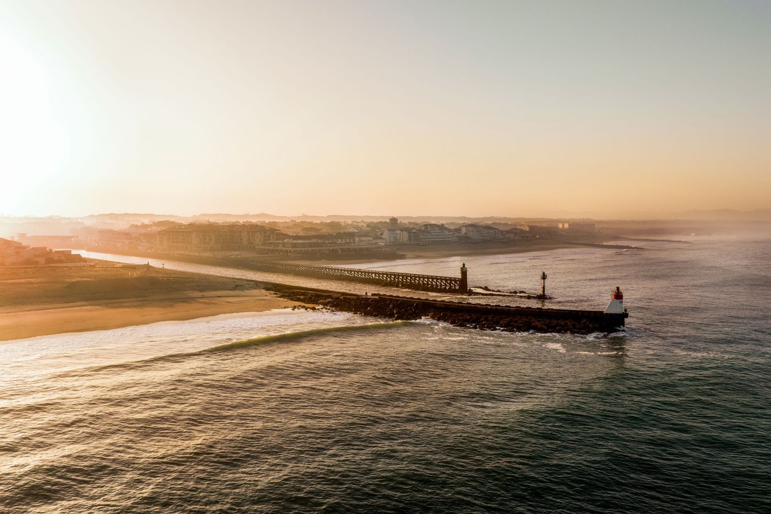 Scenic aerial view of a pier stretching into the ocean during golden sunset with waves crashing against the coastline.