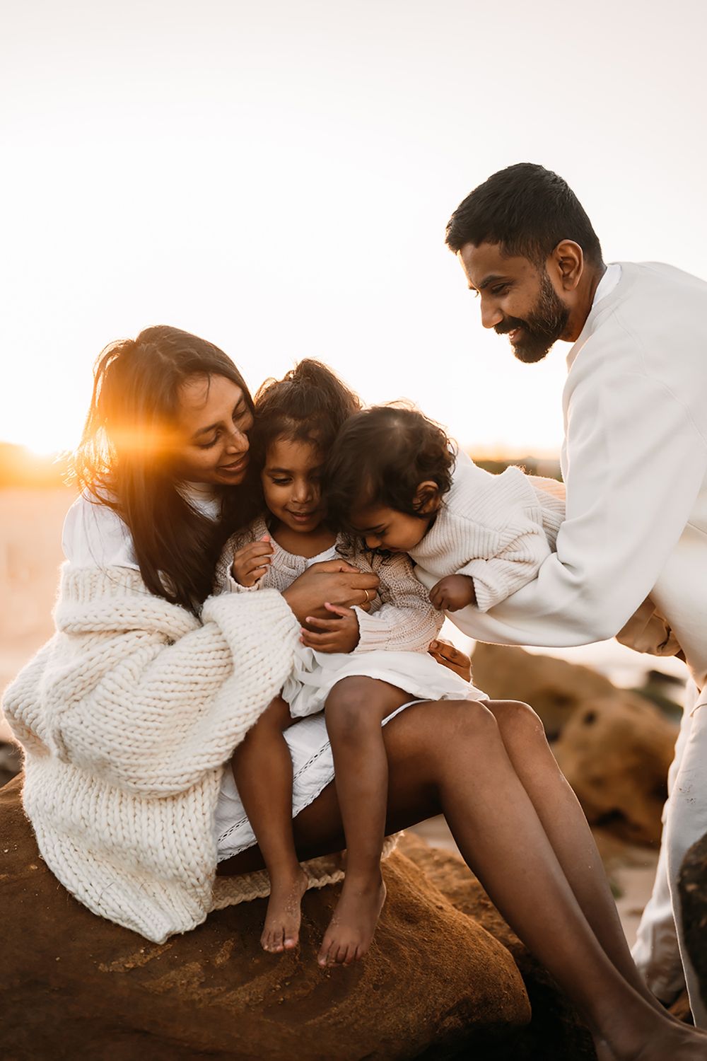 A family shares tender moments together on the beach during sunset, wearing white outfits and embracing lovingly.
