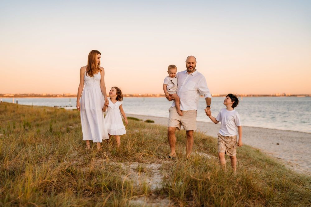 Family wearing white outfits walks along beach at sunset with ocean and golden sky in background.