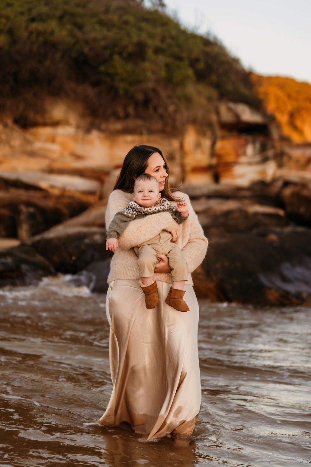 A tender moment captured at sunset on a rocky beach shore with warm golden lighting and neutral-toned clothing.