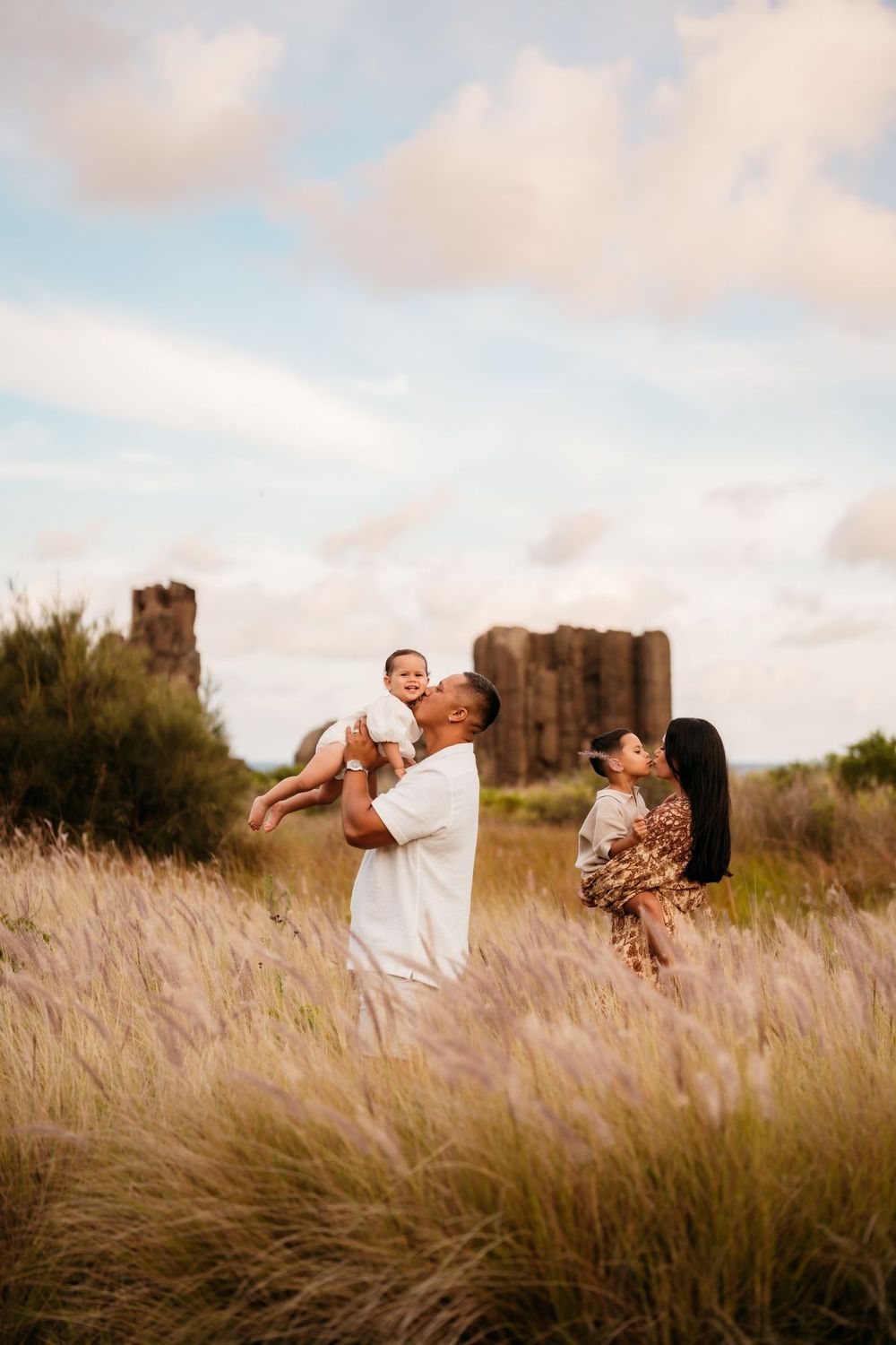 A family embraces and dances in a golden field of tall grass during sunset with large scenic rocks in the background.