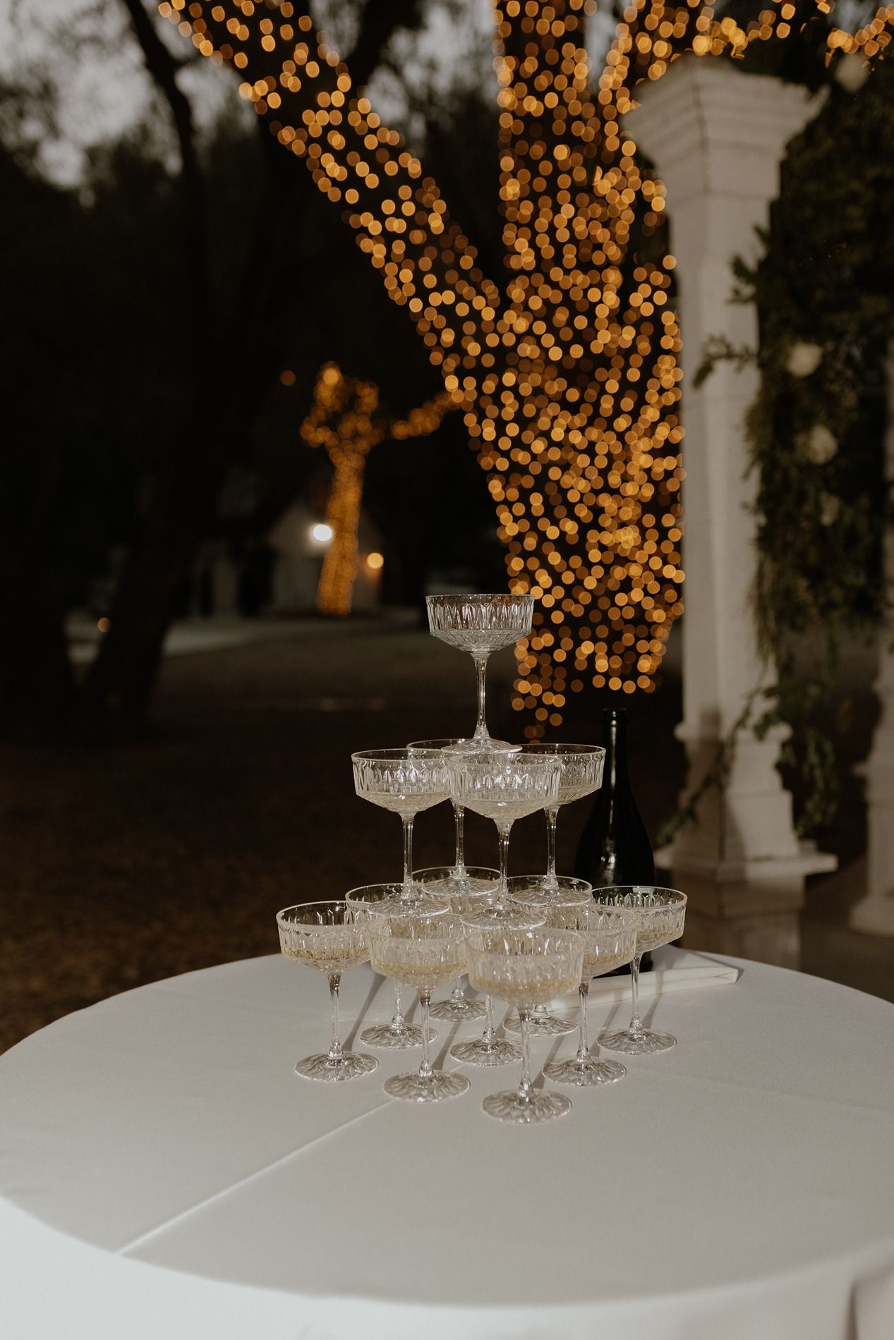 Vintage champagne coupe glasses arranged in a pyramid tower on a white table with twinkling lights in background at The Oaks at Duncan Lane.