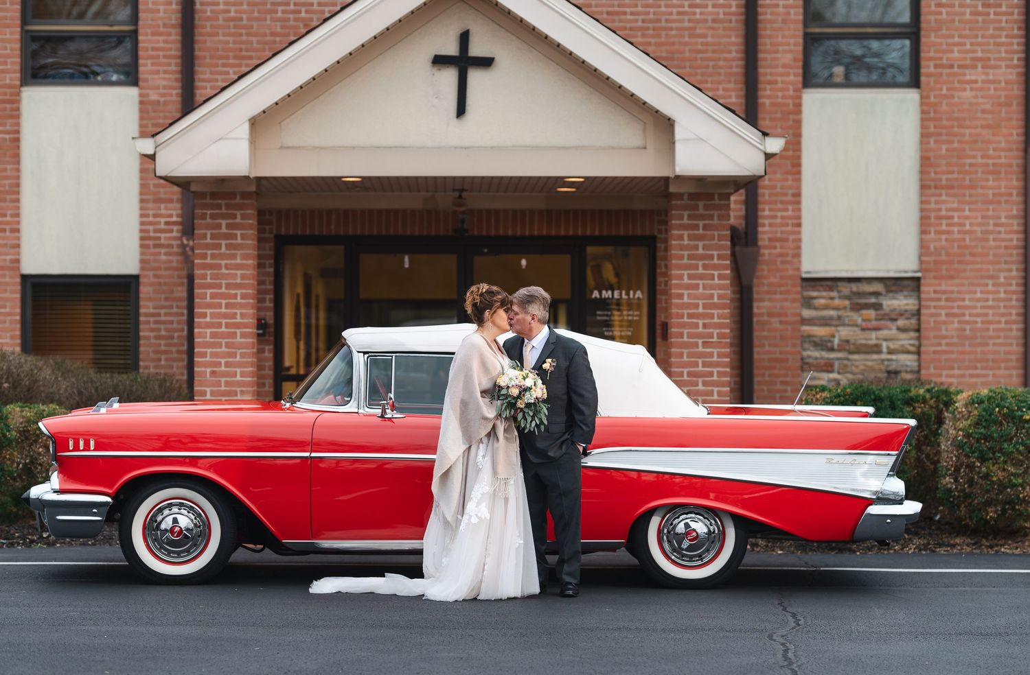 Classic red and white vintage car parked in front of brick church building during wedding ceremony.
