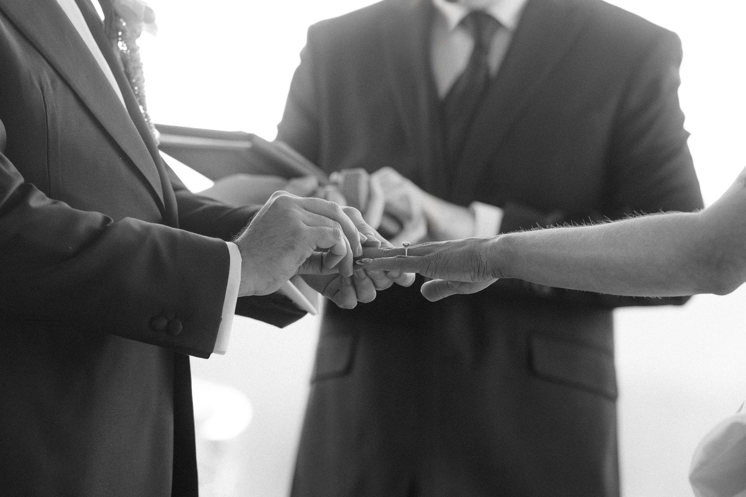 A couple holds hands during a wedding ceremony while exchanging rings in an elegant indoor setting.