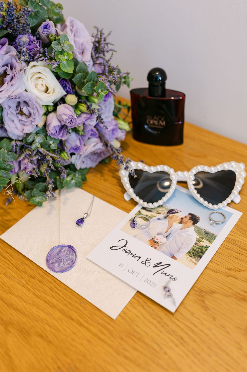 Wedding details including bouquet, invitation, rings and pearl sunglasses styled on wooden table.