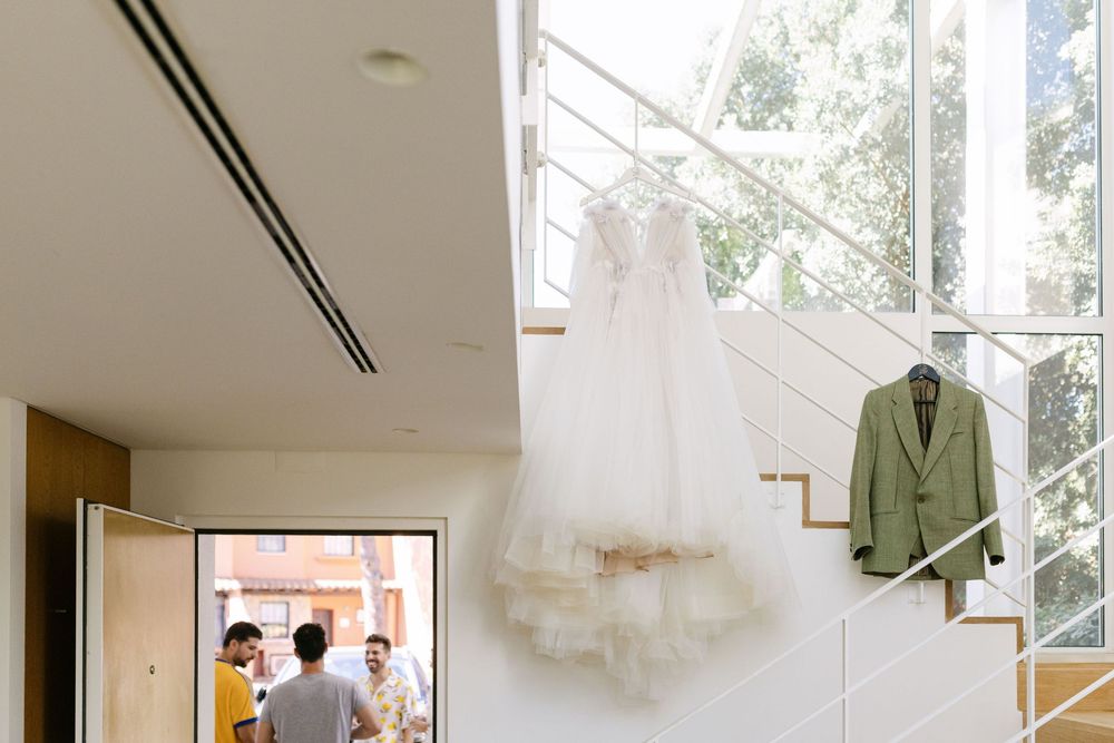 Wedding dress and green suit jacket hanging by staircase window in Lisbon wedding morning.