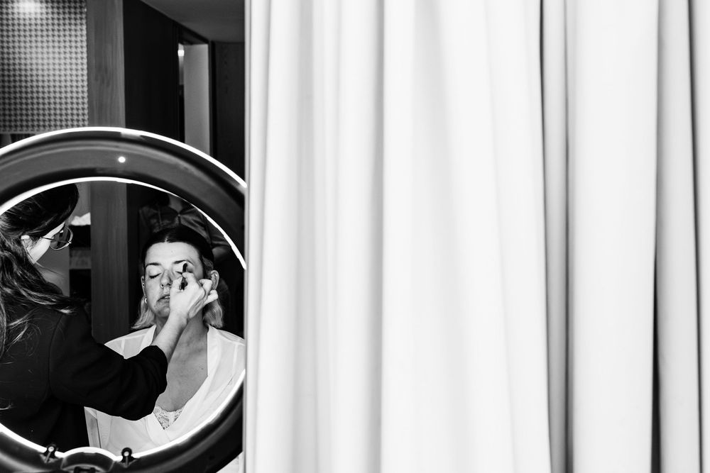 Makeup artist applying eyeshadow to bride reflected in circular mirror during getting ready.