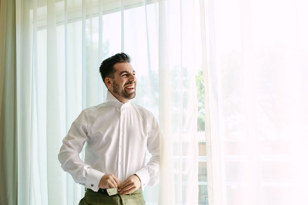 Groom buttoning shirt cuffs by window light before Forte da Crismina wedding ceremony.