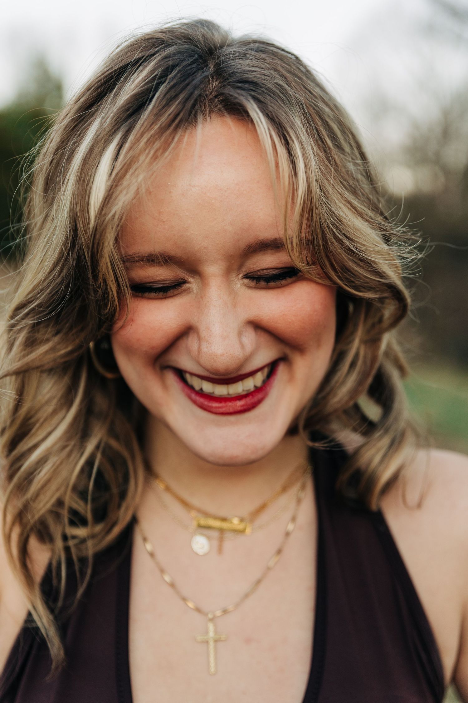 Close-up portrait of someone with wavy blonde hair wearing a brown top and layered gold necklaces, smiling outdoors.