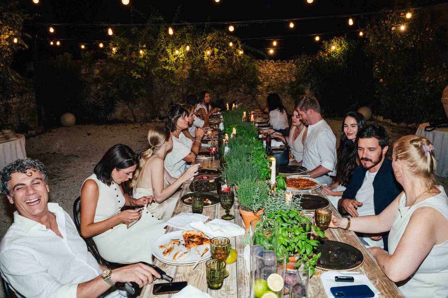 lange tafel in einem garten in italien mit Lichterketten überm tisch und mit vielen kräutern auf dem tisch dekoriert.