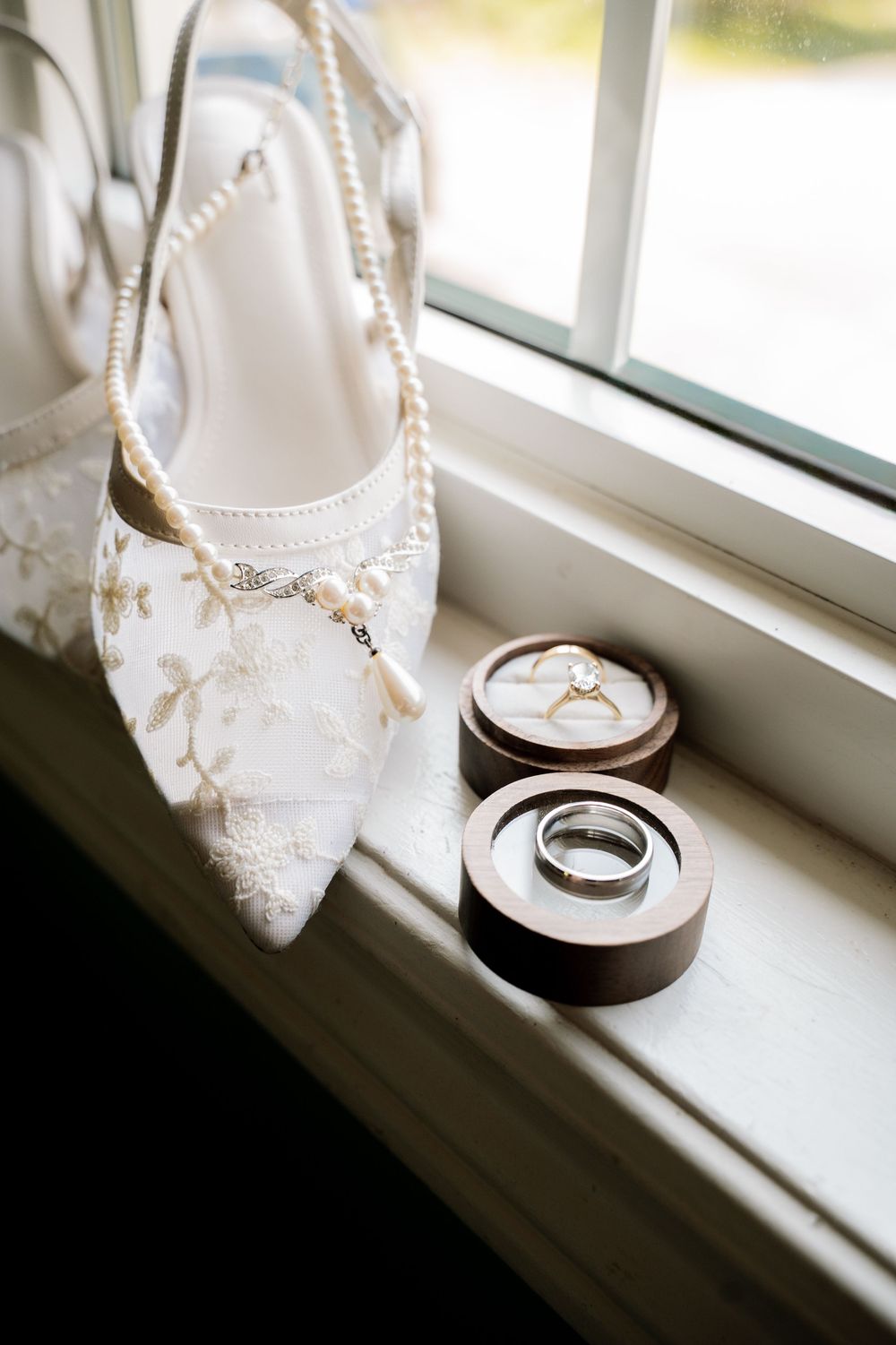 White lace bridal shoes and wedding rings displayed on a windowsill in natural light.