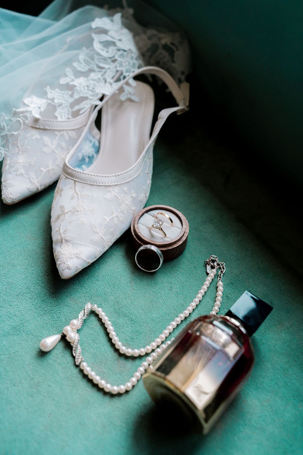White lace bridal shoes arranged with pearl necklace and perfume bottle on teal background in elegant wedding detail shots.