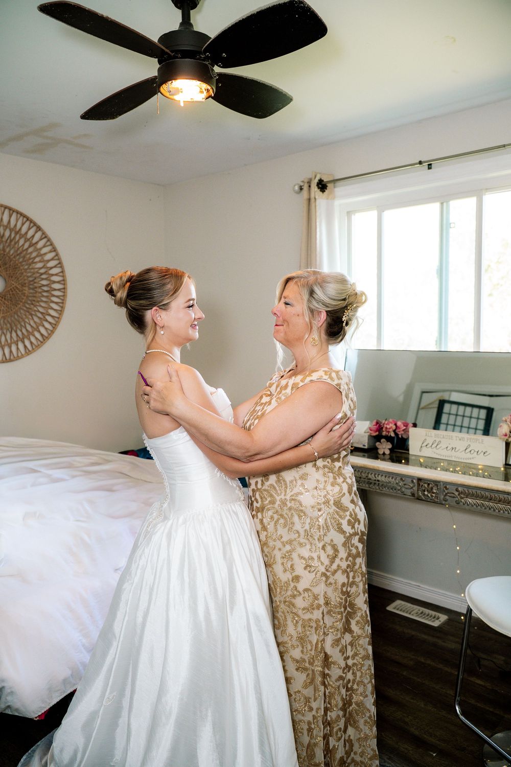Two women in formal attire share an emotional embrace during a special occasion in a sunlit room.